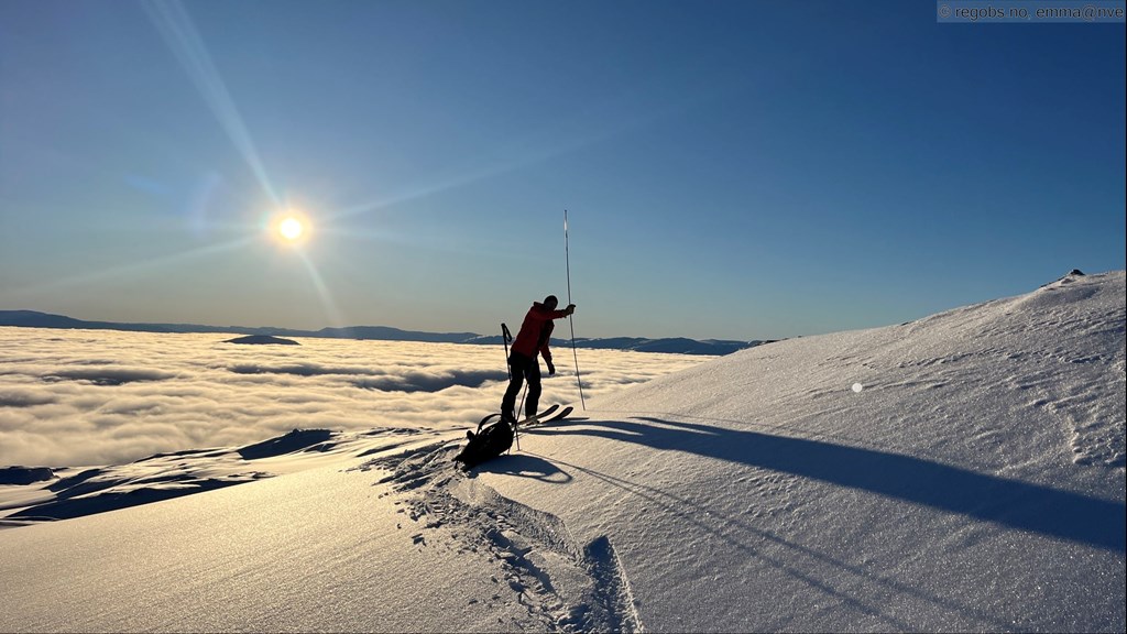 Bildet viser en som undersøker snøen i fjellet. Nede i dalen ligger det tåke. 