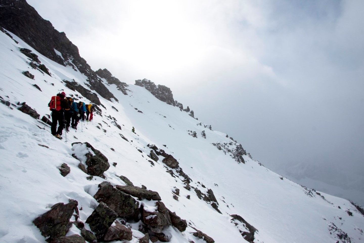 En gruppe på fottur går i en fjellside hvor det er lite snø og mye oppstikkende stein.
